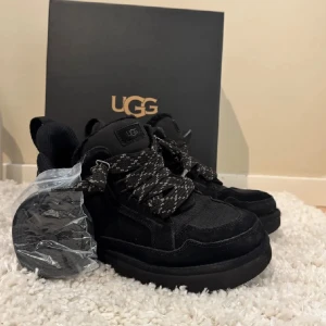 Svarta sneakers från UGG - Snygga svarta sneakers från UGG med chunky sula och grova snören i svart och vitt mönster. Skorna är tillverkade i mocka och textil, perfekta för dig som gillar en sportig och trendig look. Extra skosnören medföljer.