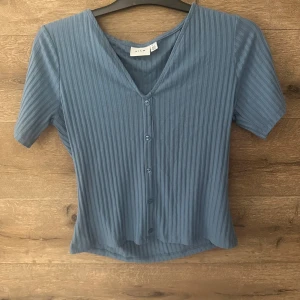 Blå ribbad blus från Vila - Säljer en blå ribbad blus från Vila med v-ringning och knappar framtill. Toppen har korta ärmar och en lätt croppad passform. Perfekt för dig som gillar en enkel men snygg look med lite struktur i tyget.