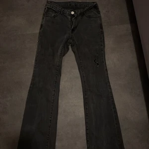 Svarta bootcut jeans med sliten detalj - Snygga svarta jeans med bootcut passform och klassisk femficksdesign. Jeansen har en sliten detalj på ena benet och stängs med knapp och dragkedja.