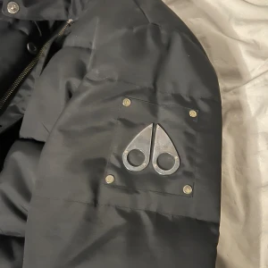 Svart dunjacka Moose Knuckles L - Säljer en svart dunjacka från Moose Knuckles i storlek L. Jackan har ikoniska metall-loggan på ärmen, broderad insida med hockeymotiv och grov dragkedja. Tillverkad i Kanada och fylld med dun för extra värme. Perfekt för kalla dagar.