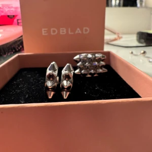 Silverset med ring och örhängen från Edblad - Coolt silverset från Edblad med en chunky ring och matchande örhängen, båda dekorerade med konformade nitar för en edgy vibe. Perfekt för dig som vill sticka ut med accessoarer som har attityd och modern design.