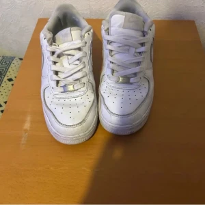 Nike Air Force 1 vita sneakers - Klassiska Nike Air Force 1 sneakers i helvitt skinn med perforerad tå, rund tå och platt sula. Skorna har snörning och den ikoniska swoosh-loggan på sidan. Perfekta för dig som gillar en clean och tidlös streetstil.