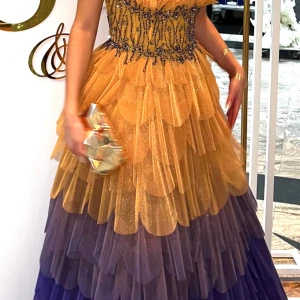 Glittrig lila och gul balklänning - Magisk långklänning med glittriga lager i gult och lila. Klänningen har en figurnära överdel med dekorativa pärlor och volangliknande kjol i flera lager. Perfekt för bal eller fest där du vill sticka ut.