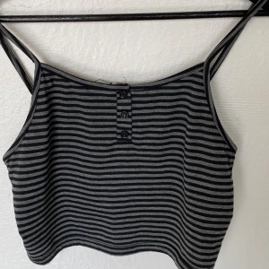 Randig croppad linnetopp från Romwe - Säljer en croppad linnetopp från Romwe i svart och grått med smala axelband. Toppen har horisontella ränder och tre knappar framtill som detalj. Perfekt för varma dagar eller att styla lager på lager.