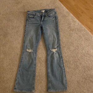 Bootcut jeans från Gina Tricot, ljusblå - Ljusblå jeans från Gina Tricot i bootcut-modell med slitna detaljer på knäna. Klassisk femficksdesign och knappstängning. Jeansen har en avslappnad vibe och är perfekta för dig som gillar en lite retro look.