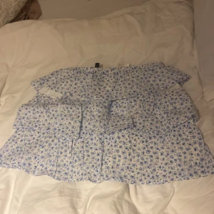 Blommig volangkjol från H&M Divided XS - Supersöt kort kjol från H&M Divided i storlek XS. Kjolen är vit med blått blommigt mönster och har flera volanger som ger en flowy och feminin look. Perfekt för varma dagar och har en elastisk midja för skön passform.