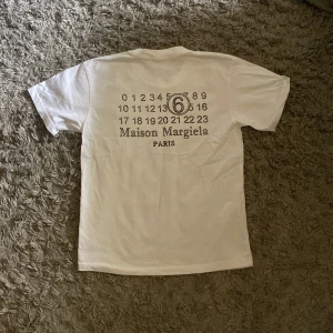 Maison Margiela vit t-shirt med print - Vit t-shirt från Maison Margiela med ikoniskt siffertryck och logga på ryggen. Gjord i mjuk bomull och är i storlek M. 