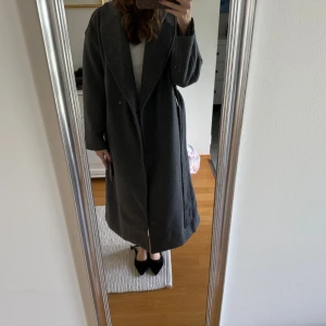 Mörkgrå lång kappa med bälte - Säljer en snygg mörkgrå kappa i lång modell med bred krage och knytskärp i midjan. Jackan har raka ärmar, stora fickor och en clean look som passar perfekt till höst och vinter. Materialet känns mjukt och värmande, troligen ullblandning.