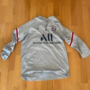 Grå PSG långärmad fotbollströja - Snygg grå långärmad PSG-tröja från Jordan med svarta prickar, klubbmärke på bröstet och Accor Live Limitless-tryck. Röd, vit och blå rand på ärmarna. Materialet är lätt och andas, perfekt för träning eller match.