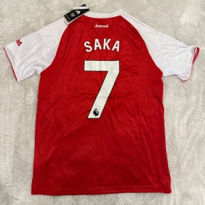 Arsenal Saka 7 matchtröja Adidas - Arsenal fotbollströja med Saka 7 på ryggen. Röd kropp med vita ärmar och vita detaljer, broderad klubbmärke och Adidas-logga. Materialet är lätt och andas, perfekt för match eller träning. Klassisk design med sponsortryck och Premier League-märke på ärmen.