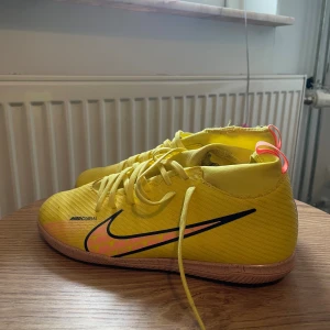 Nike Mercurial gula fotbollsskor - Snygga Nike Mercurial fotbollsskor i en riktigt poppig gul färg med rosa och svarta detaljer. Skorna har snörning, platt sula och är tillverkade i syntetmaterial. Perfekta för dig som vill sticka ut på planen med stilrena och sportiga skor.