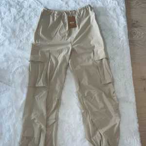 Beige cargopants från Aimn - Snygga beige cargopants från Aimn med elastisk midja och stora fickor på sidorna. Byxorna har en loose passform och är tillverkade i ett lätt syntetmaterial, perfekta för en avslappnad streetstil.
