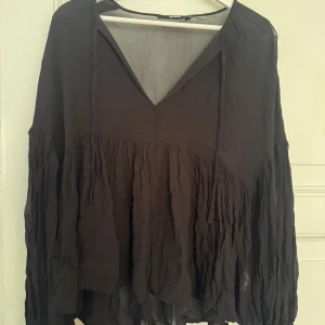 Svart blus från Bik Bok  - Luftig svart blus från Bik Bok i storlek XS. Blusen har v-ringning med knytband, långa ärmar och volangig, rynkad nederdel. Tunt, flowy material som ger en avslappnad boho-vibe. Perfekt att styla med jeans eller kjol för en cool look.