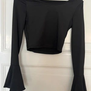 Svart croppad topp med trumpetärmar - Svart croppad topp från SHEIN med långa trumpetärmar som ger en snygg statement-look. Toppens material är mjukt och stretchigt, perfekt för att skapa en cool outfit. Enkel rund halsringning och figurnära passform.