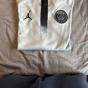 PSG x Jordan vit fotbollströja - Snygg vit PSG fotbollströja i samarbete med Jordan. Tröjan har svart vertikal rand i mitten, PSG-logga på bröstet och klassiska Jumpman-loggan. Tillverkad i lätt och ventilerande material, perfekt för träning eller match. Svarta detaljer vid ärmslut.