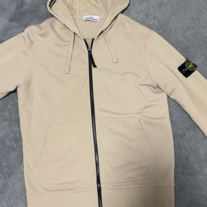 Beige hoodiejacka från Stone Island - Snygg beige hoodiejacka från Stone Island med klassisk patch på ärmen. Jackan har huva med snörning, stor magficka och svart dragkedja framtill. Perfekt för dig som gillar streetwear och vill ha en clean look.