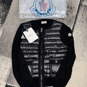 Svart quiltad cardigan jacka Moncler - Svart cardigan jacka från Moncler med quiltad, glansig front och två fickor med knappdetaljer. Jackan har långärmade stickade ärmar och dragkedja framtill. Moncler-logga på ärmen och rund halsringning ger en clean och exklusiv look.❤️
