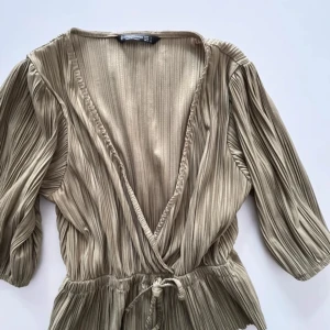 Topp  - Säljer en beige omlottopp från Stradivarius med snygg ribbad struktur och knyt i midjan. Toppen har trekvartsärm och djup v-ringning som ger en chill och trendig vibe. Perfekt att styla med jeans eller kjol för en cool look.