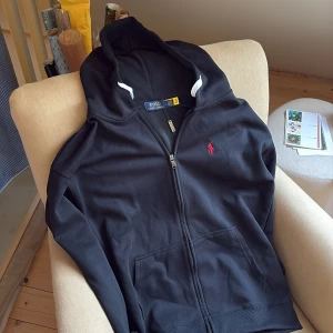 Svart hoodie från Polo Ralph Lauren - Svart hoodie från Polo Ralph Lauren med dragkedja och vit snörning i huvan. Klassisk röd logobrodering på bröstet. . Snygg och enkel passar till det mesta.