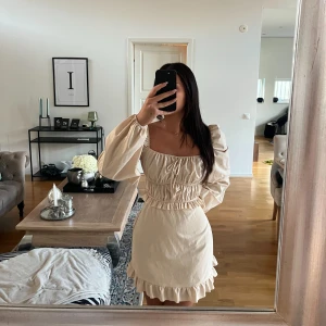 Beige klänning  - Säljer en beige kort klänning med puffiga långärmar och volangdetaljer vid nederkanten. Klänningen har en fyrkantig urringning och smockad midja som ger en snygg siluett. Perfekt för dig som gillar romantisk och trendig stil.