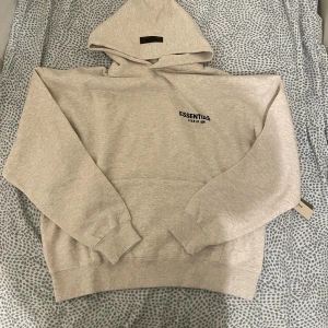 Ljusgrå Essentials Fear of God hoodie - Säljer en ljusgrå hoodie från Essentials Fear of God i storlek M. Tröjan har stor svart logga på ryggen och mindre logga på bröstet, samt huva och klassisk känguruficka. Mjuk bomullskänsla och ribbade muddar vid ärmar och nederkant. Perfekt för chill och streetwear.
