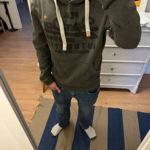 Mörkgrå Superdry hoodie med tryck - Snygg mörkgrå hoodie från Superdry med stort svart tryck på bröstet och japanska tecken. Tröjan har huva med grova vita snören och en klassisk magficka. Perfekt för dig som gillar streetwear och vill ha en chill look. Hör av dig gällande frågor, inga defekter på tröjan riktigt fint skick. Storlek M