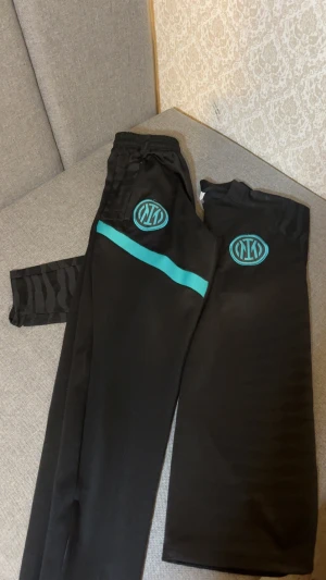 Nike Inter Milan svart träningsset - Snyggt svart träningsset från Nike och Inter Milan med turkosa detaljer och loggor. Tröjan har lång ärm, half zip och Dri-Fit ADV-tyg som andas. Byxorna har elastisk midja och matchande turkos rand. Perfekt för fotboll eller chill.