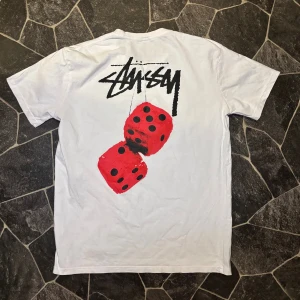  Stüssy t-shirt  - Vit t-shirt från Stüssy med klassisk logga på bröstet och stor grafisk print med röda tärningar på ryggen. Skön passform och tillverkad i mjuk bomull. Perfekt för dig som gillar streetwear och vill sticka ut med coola detaljer.