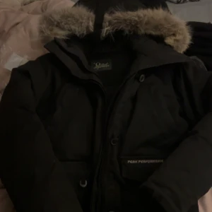Svart parkas Peak Performance XS - Svart vintage Peak Performance med huva och snygg fuskpäls runt kanten. Jackan har dragkedja och knappar framtill samt praktiska fickor. Perfekt för kalla dagar och riktigt skön passform.