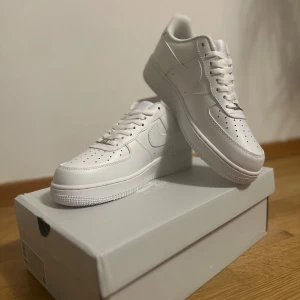 Nike Air Force 1 '07 helvita sneakers - Nike Air Force 1 '07 i helvitt skinn med klassisk siluett, perforerad tåbox och vit sula. Snygga detaljer som metallbricka med AF1-logga och ikonisk swoosh på sidorna. Perfekta sneakers för dig som gillar clean och tidlös stil.