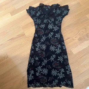 Svart blommig långklänning Eclipse - Supersnygg svart långklänning från Eclipse med blommigt mönster i vitt och röda detaljer. Klänningen har korta ärmar med spetskant och v-ringad hals med volang. Materialet är lätt och känns som polyester, perfekt för dig som gillar romantisk stil.