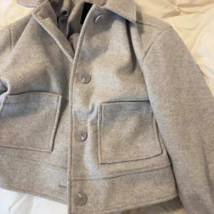 Grå ulljacka från 157, XS - Snygg grå jacka från 157 i ullmaterial, med klassisk krage och stora framfickor. Jackan har knappar framtill och på ärmsluten, samt en rak och lite croppad passform. Perfekt för dig som gillar stilrena och enkla plagg med en modern touch.