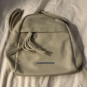 Beige ryggsäck från Anna Field - Snygg beige ryggsäck från Anna Field i mjukt syntetmaterial. Väskan har silverfärgade detaljer, dekorativ tofs framtill och dragkedja upptill. Insidan är svartfodrad med innerfack för småprylar. Perfekt storlek för vardagens alla grejer.