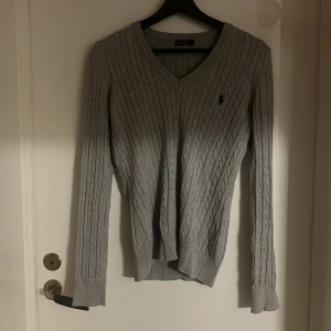 Grå kabelstickad tröja Polo Ralph Lauren - Snygg grå kabelstickad tröja från Polo Ralph Lauren med v-ringning och broderad logga på bröstet. Tröjan har lång ärm och ribbade muddar vid ärmslut och nederkant. Perfekt för dig som gillar klassisk stil med en modern touch.