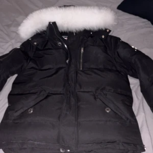 Svart dunjacka Moose Knuckles - Svart dunjacka från Moose Knuckles med vit pälskant på huvan och silverfärgade detaljer. Jackan har dragkedja, knappar och flera fickor samt logotyp på ärmen. Perfekt för kalla dagar och har en klassisk, sportig look.