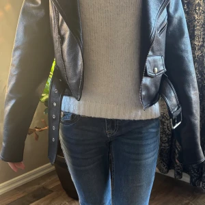 Svart skinnjacka med bälte och dragkedja - Snygg svart skinnjacka med klassisk bikerstil, detaljerad med silvriga dragkedjor, tryckknappar och ett justerbart bälte i midjan. Jackan har breda slag och fickor med lock framtill. Använd fåtal gånger. 