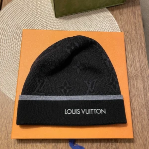 Svart Louis Vuitton mössa med logga - Exklusiv svart mössa från Louis Vuitton med diskret monogrammönster och grå rand längs kanten. Märkets logga är broderad framtill. Mössan är stickad och tillverkad i Italien, perfekt för dig som vill ha en lyxig och stilren accessoar.