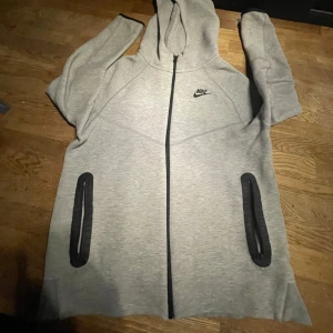 Grå hoodie från Nike XL - Snygg grå hoodie från Nike i storlek XL. Tröjan har hel dragkedja framtill, två svarta fickor med dragkedja och en klassisk huva. Nike-logga på bröstet. Perfekt för chill eller träning, mjukt material och långärmad design. Priset kan diskuteras, tröjan är äkta.