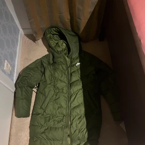 Grön lång pufferjacka från Nike - Säljer en grön pufferjacka från Nike med huva och dragkedja framtill. Jackan har ett quiltat mönster och är perfekt för kalla dagar. Nike-loggan syns tydligt på bröstet. Jackan är lång i modellen och har en clean, sportig look.