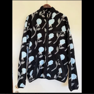 Streetwear Blommig fleecejacka från HUF - Säljer en svart fleecejacka från HUF med ljusblå blommor och grå blad över hela jackan. Jackan har hel dragkedja framtill, två sidofickor med dragkedja och en bröstficka med HUF-logga. Perfekt för dig som gillar streetwear med unik design.