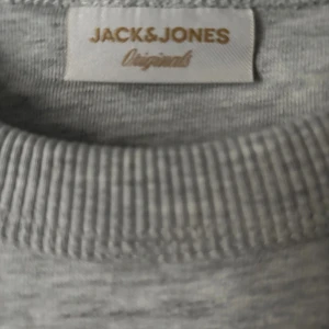 Grå sweatshirt från Jack & Jones - Snygg ljusgrå sweatshirt från Jack & Jones Originals med svart broderad text 'Originals Studio' på bröstet. Tröjan har rund hals, ribbade muddar vid ärmslut och nederkant samt lång ärm. Perfekt för chill dagar och enkel att matcha med jeans.