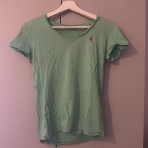 Mintgrön t-shirt från Ralph Lauren - Fräsch mintgrön t-shirt från Ralph Lauren med v-ringad hals och korta ärmar. Klassisk rosa broderad logga på bröstet. Skön och lätt i bomull, perfekt för en chill stil. Passar dig som gillar enkel design med en touch av färg.