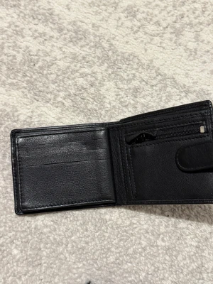 Svart skinnplånbok från Pierre Cardine ej använd  - Snygg svart plånbok i äkta skinn från Pierre Cardin. Plånboken har flera kortfack, myntfack med dragkedja och genomskinliga fack för ID eller bilder. Stilren och praktisk design som passar perfekt i fickan.