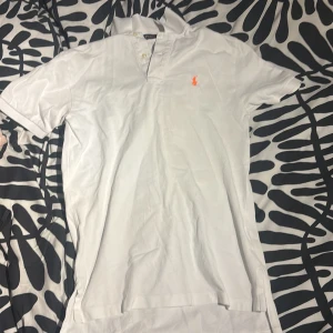 Vit pikétröja från Polo Ralph Lauren - Vit pikétröja från Polo Ralph Lauren i storlek S. Klassisk design med krage, två knappar och orange broderad logga på bröstet. Tröjan är gjord i mjuk bomull och har korta ärmar. Perfekt för en clean och fräsch look.
