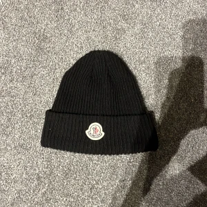 Svart ribbad Moncler mössa - Svart ribbstickad mössa från Moncler med klassisk uppvikt kant och ikonisk Moncler-logga framtill. Snygg och enkel design som passar perfekt till kalla dagar. Materialet är mjukt och värmande, och mössan har en stilren look som funkar till alla outfits.
