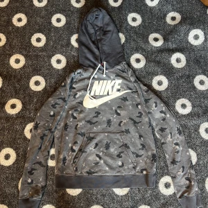 Grå camo Nike hoodie med huva - Säljer en grå hoodie från Nike med camouflagemönster och stor vit logga på bröstet. Tröjan har huva med snörning och en klassisk magficka. Perfekt för dig som gillar streetwear och sportig stil.