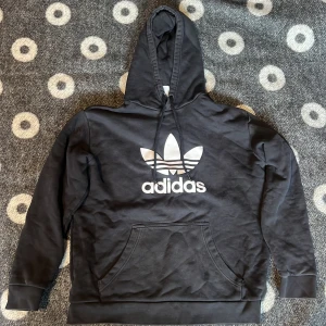 Svart Adidas hoodie  - Svart hoodie från Adidas med stor vit trefoil-logga och text på bröstet. Tröjan har huva med snörning och en klassisk magficka. Perfekt för dig som gillar sportig och avslappnad stil.