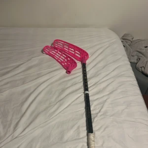 Salming Trident innebandyklubba - Salming Trident innebandyklubba med två rosa blad. Skaftet är svart med guldtext och har viss kosmetisk slitage, men är i gott skick. Perfekt för både träning och match. Greppet ser fräscht ut och bladen är hela utan sprickor. Scick 9/10 då knappt använd 29 flex men kapad till 97. Priset är ej hugget i sten