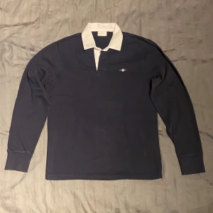 Gant rugger rugbytröja  - Marinblå rugbytröja från Gant. Tidlöst plagg men en stilren enfärgad design med vit krage i kontrast. Muddar vid handleder och muddlös i nedkant