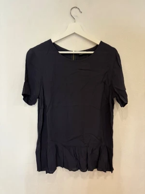Mörkblå blus med volang från KappAhl - Snygg mörkblå blus från KappAhl i storlek M. Blusen har rund halsringning, korta ärmar och en guldfärgad dragkedja i ryggen. Nedtill finns en bred volang som ger en cool och feminin touch. Perfekt för dig som gillar stilrena detaljer.